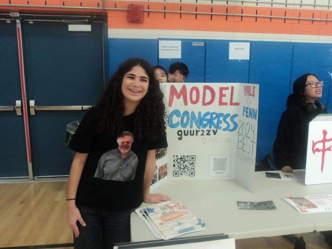 A student stands smiling beside a club table in a school gymnasium. Behind the table a hand-lettered tri-fold board reads MODEL CONGRESS in red and blue marker, with a class code and two QR codes below it; printed leaflets are stacked on the table. The student wears a black T-shirt printed with a photograph of a bearded man in a checked shirt. Other club tables are visible further down the row.