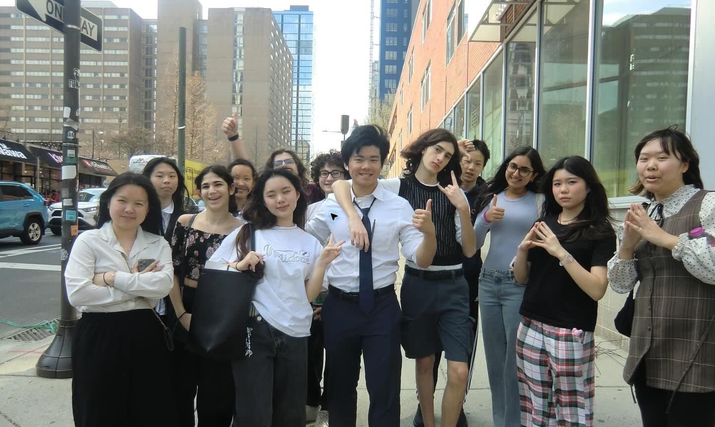Twelve students stand shoulder to shoulder on a city sidewalk beneath a one-way sign, several giving thumbs-up or peace signs. They are dressed in a mix of formal and everyday clothes; apartment towers, a red brick building and a glass storefront rise behind them.