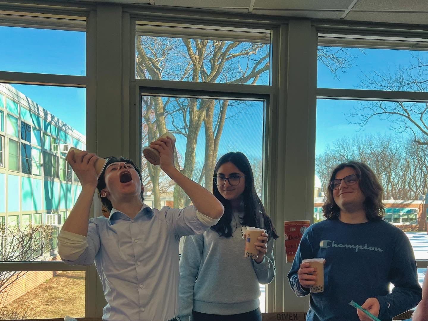 Three students stand behind a table indoors, each holding a clear cup of milk tea with tapioca pearls. One tips his cup high toward his open mouth while the other two face the camera; tall sunlit windows and bare winter trees fill the wall behind them, and packets of crisps are stacked along the table in front.