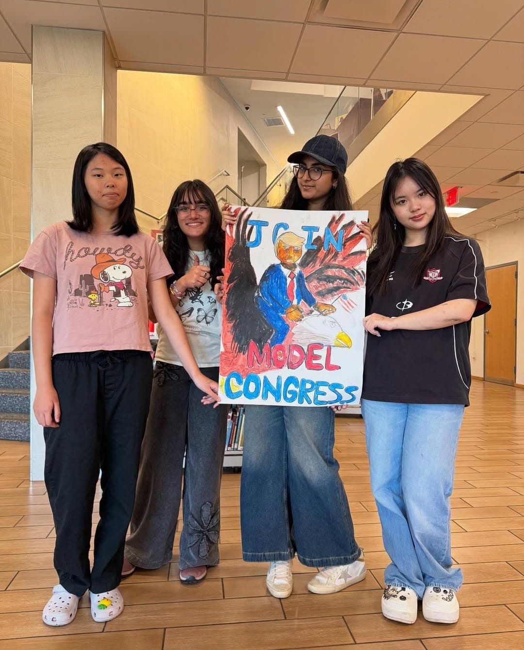 Four students stand together in a school lobby with a staircase behind them. The two in the middle hold up a large hand-painted poster reading JOIN MODEL CONGRESS in blue and red block letters, above a painted eagle and a figure in a blue suit.