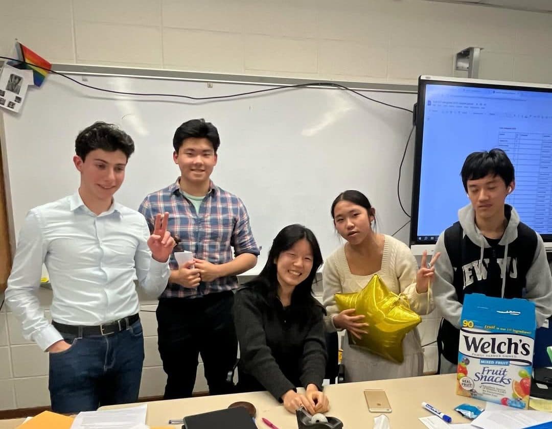Five students at the head of a long classroom table, one seated and four standing behind her, two of them making peace signs. The table in front of them is covered with printed documents, pens, a tape dispenser and a small wooden block; a whiteboard and a wall-mounted display screen fill the wall behind.